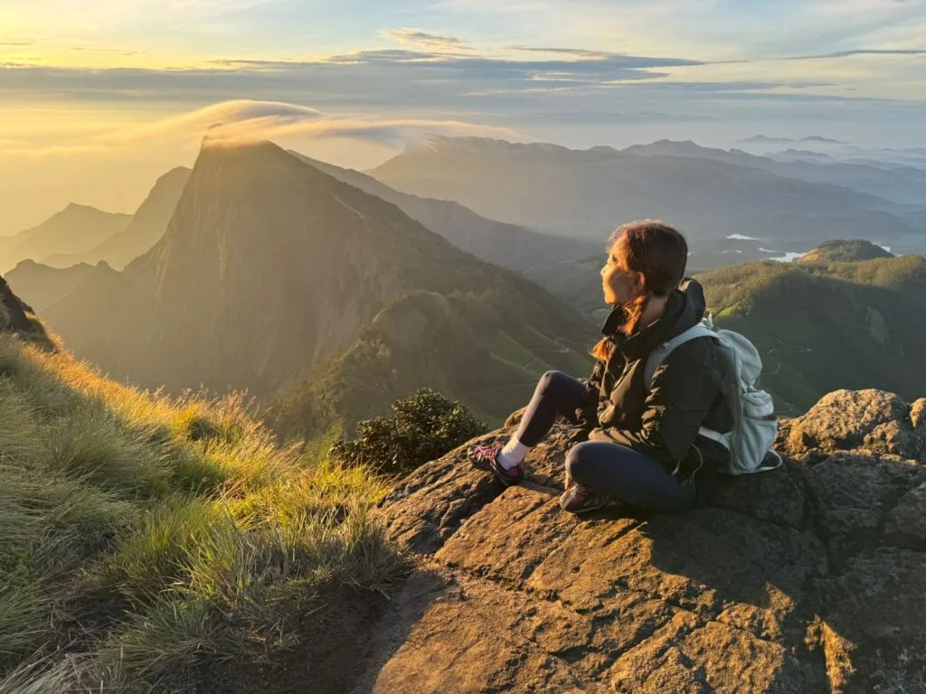 A guest sits on a grassy ridge in the Western Ghats, watching the sun rise over layers of misty hills