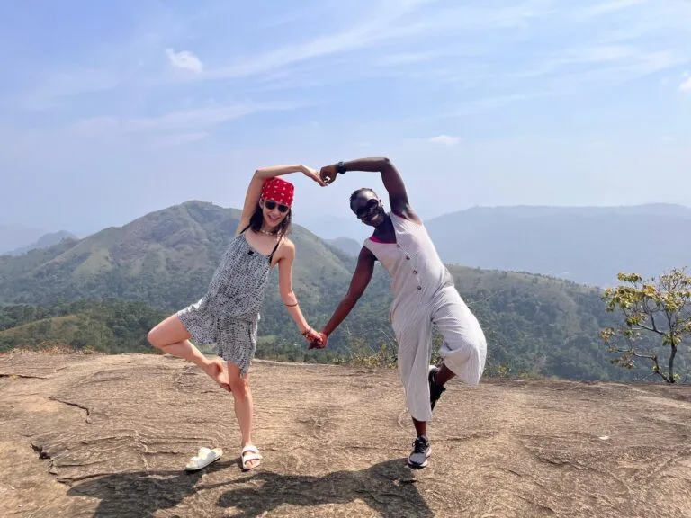 Two guests balance in tree pose and make a heart with their arms on a hilltop