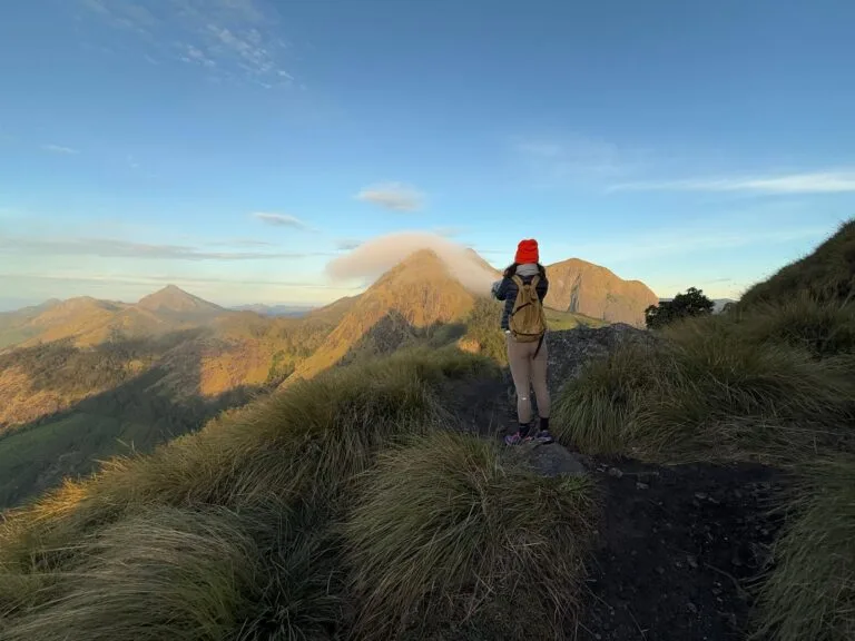 A trekker stands on a grassy ridge with a pointed peak behind them