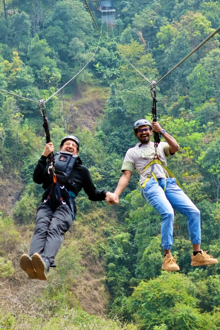 Two guests in harnesses laugh as they ride a zipline above the forest